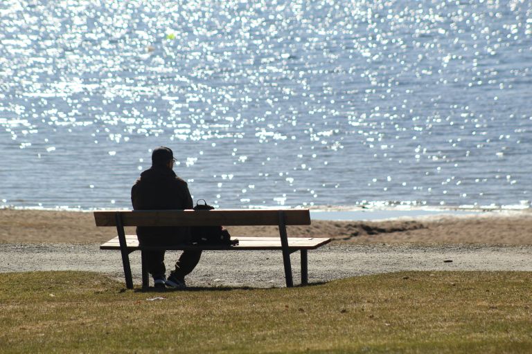 A young adult sitting alone on a park bench while people interact in the background, symbolizing loneliness and social isolation.